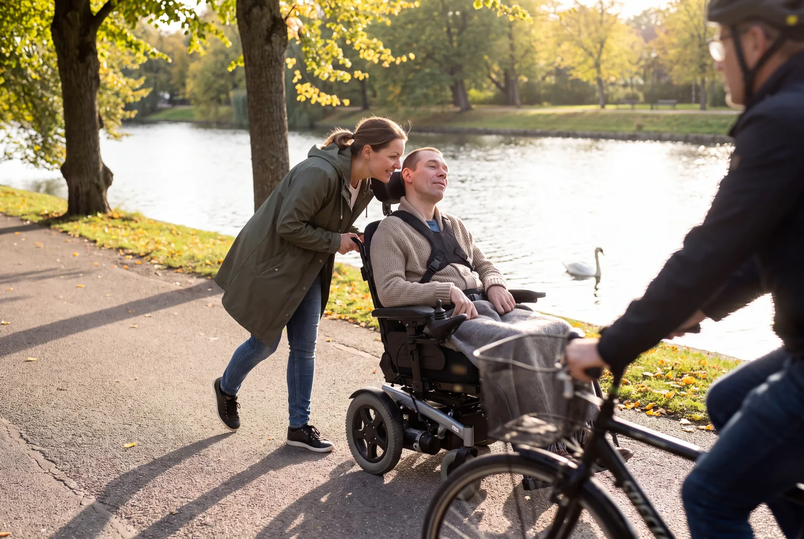 En assistent kör en man i permobil längs kanalen i Slottsparken i Malmö en solig höstdag, en cyklist passerar i förgrunden.