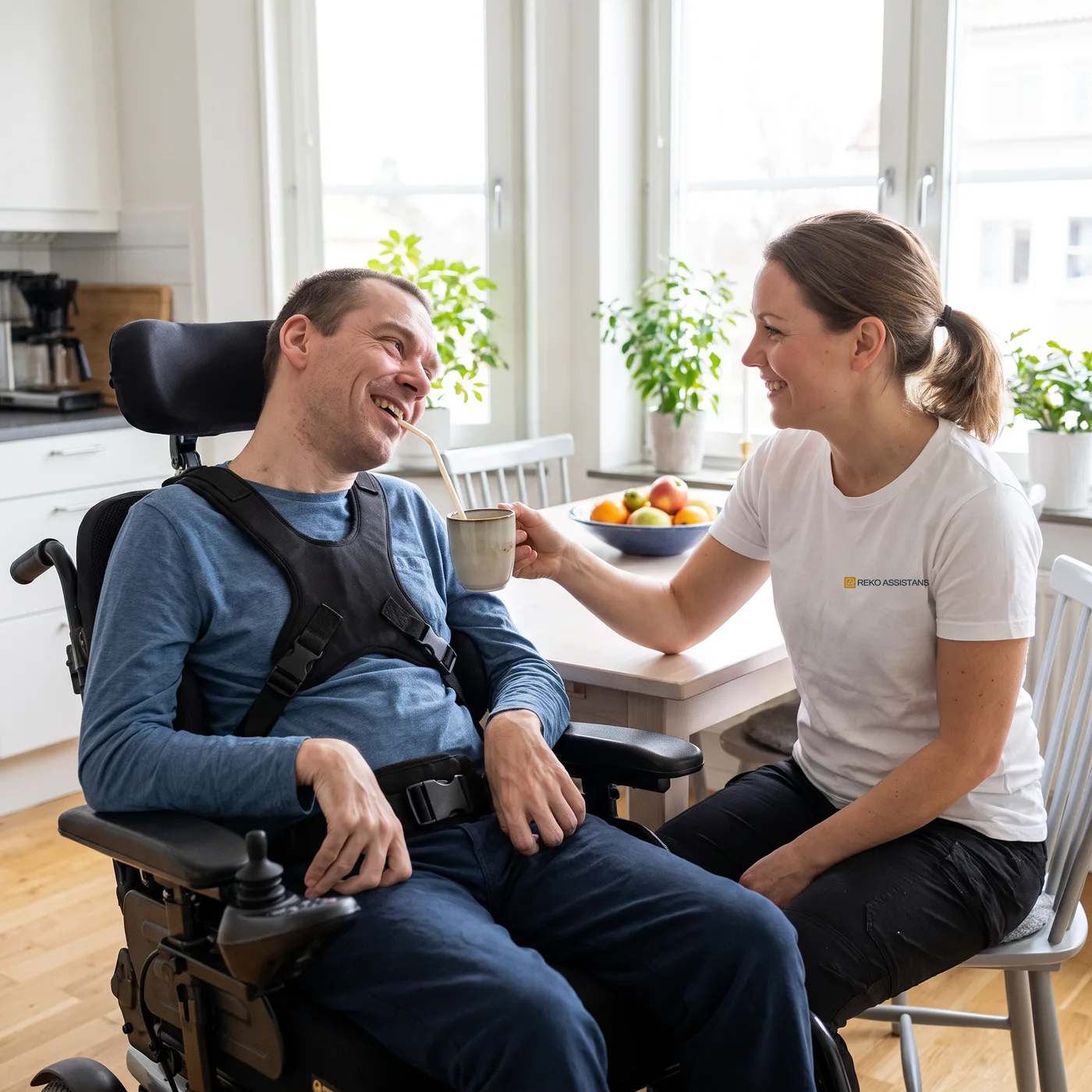 En personlig assistent håller fram en mugg med sugrör åt en man i permobil vid ett köksbord.